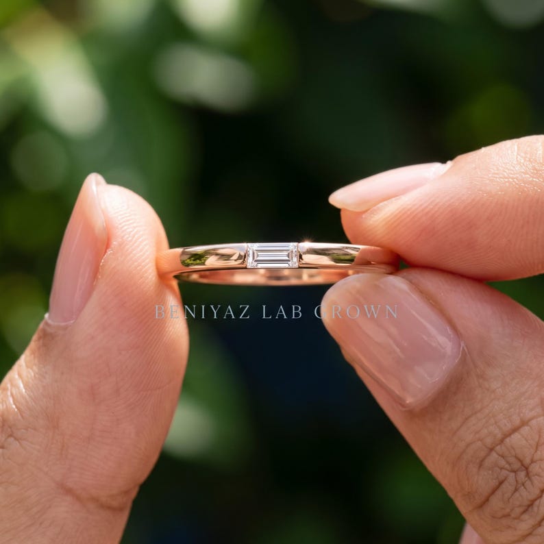 Close up hand held east west baguette lab grown diamond ring with a minimalist polished gold wedding band.