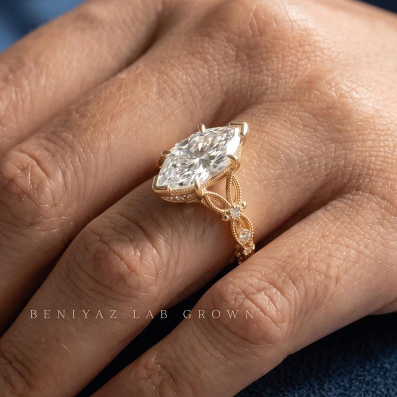 May include: A marquise-cut diamond ring set in a gold band with intricate filigree detailing and small accent stones along the shank. The text BENIYAZ LAB GROWN is visible at the bottom of the image, featuring a clear, faceted center stone.