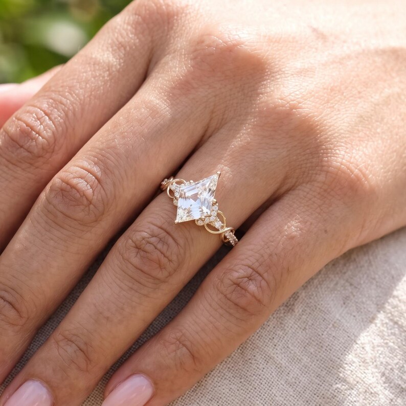 A woman's hand with pale pink manicured nails wearing a kite cut lab grown diamond engagement ring over cream knitted fabric. The large geometric center diamond and ornate pavé band are clearly visible, showing how the ring looks when worn.
