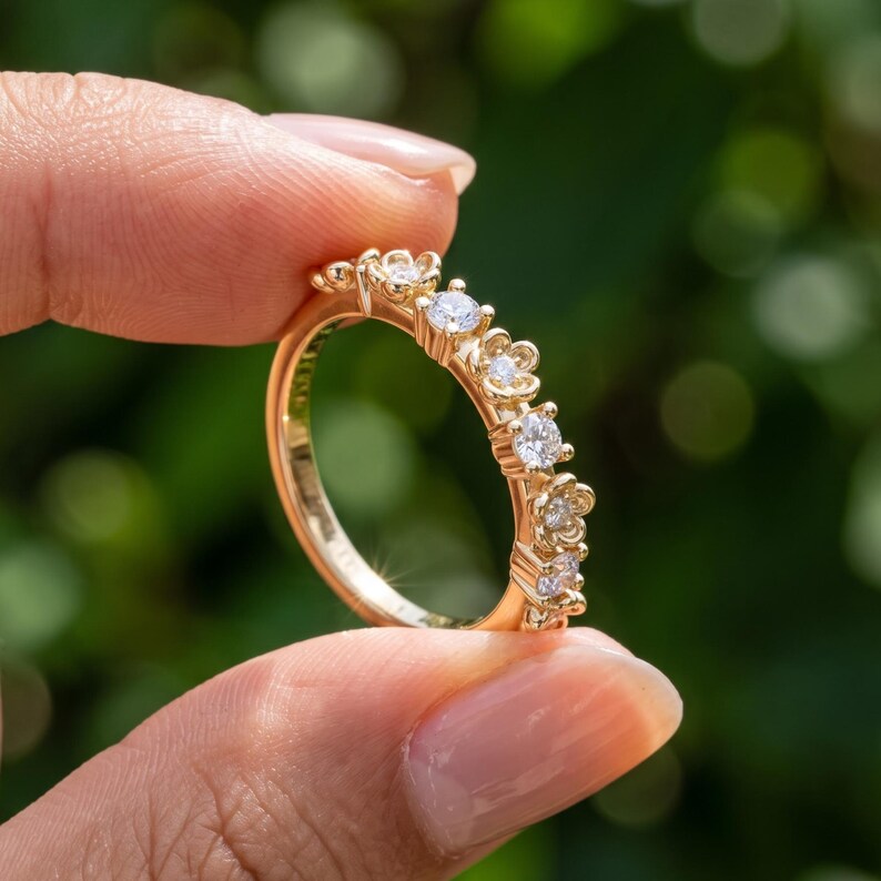 May include: A gold ring featuring a series of small, flower-shaped settings along the band, each containing a single round, clear gemstone. The metal has a polished, warm yellow tone, and the floral design elements are arranged in a row.