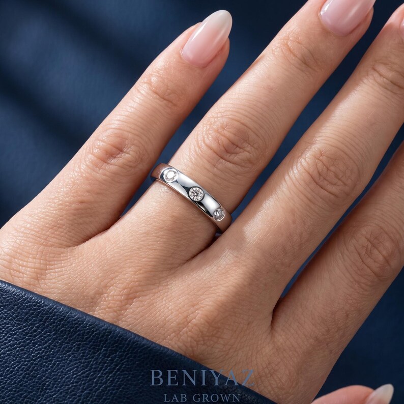 May include: A polished silver-toned band ring featuring three round, bezel-set clear stones embedded along the top surface. The ring is worn on a finger, and the text BENIYAZ LAB GROWN is visible at the bottom of the image in a dark blue font.