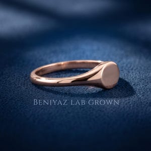 Rose gold tone round signet ring on blue background with plain circular face, tapered shank and slim half round band