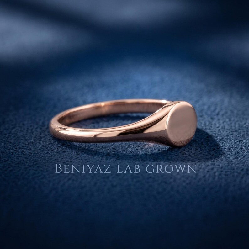 Rose gold tone round signet ring on blue background with plain circular face, tapered shank and slim half round band