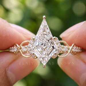 Detailed frontal macro of a kite shaped lab grown diamond engagement ring held between fingers. The angular diamond center is complemented by small sparkling diamonds along an elaborate pavé setting, with the ring photographed against a softly blurred green background.