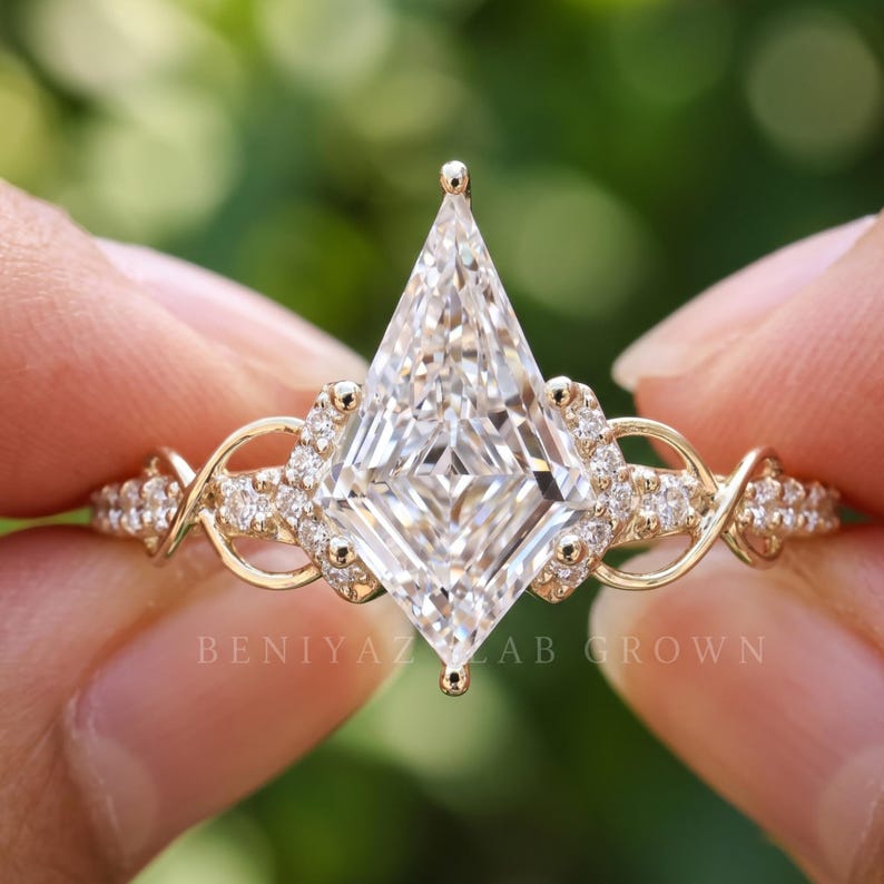 Detailed frontal macro of a kite shaped lab grown diamond engagement ring held between fingers. The angular diamond center is complemented by small sparkling diamonds along an elaborate pavé setting, with the ring photographed against a softly blurred green background.