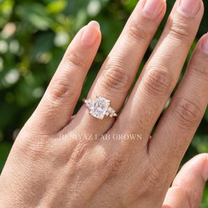 May include: A rose gold ring featuring a large rectangular radiant-cut center stone flanked by three smaller round stones on each side. The band is thin and metallic. The text BENIYAZ LAB GROWN is visible on the lower portion of the image.
