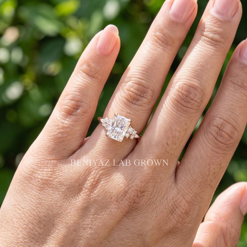 May include: A rose gold ring featuring a large rectangular radiant-cut center stone flanked by three smaller round stones on each side. The band is thin and metallic. The text BENIYAZ LAB GROWN is visible on the lower portion of the image.