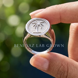 Hand holding a sterling silver palm tree signet ring showing the round medallion profile, smooth tapered shoulders and polished band