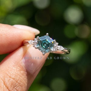 Blue moissanite engagement ring held between fingers, showing full hexagon center stone and symmetrical cluster side design.
