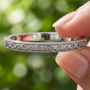 May include: A silver-toned metal ring featuring an intricate engraved scrollwork pattern along the band, bordered by delicate beaded edges. The text BENIYAZ LAB GROWN is printed in small, clear letters on the surface of the ring band.