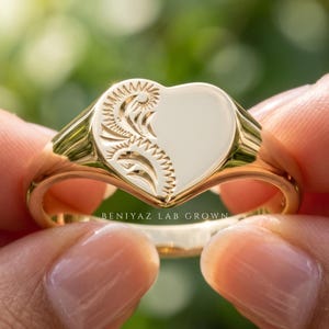 Ladies heart signet ring held between fingers with ornate floral and scroll engraving, smooth polished heart face and tapered shoulders