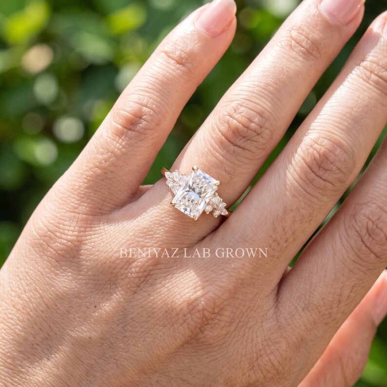 May include: A gold ring featuring a large rectangular radiant-cut center stone flanked by smaller cluster stones on each side. The text BENIYAZ LAB GROWN is visible on the image. The ring has a thin gold band and a prong setting for the stones.