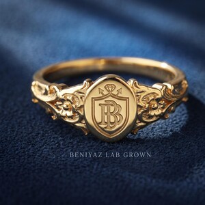 Custom monogram oval signet ring in yellow gold with an engraved crest design and ornate floral scrollwork shoulders.