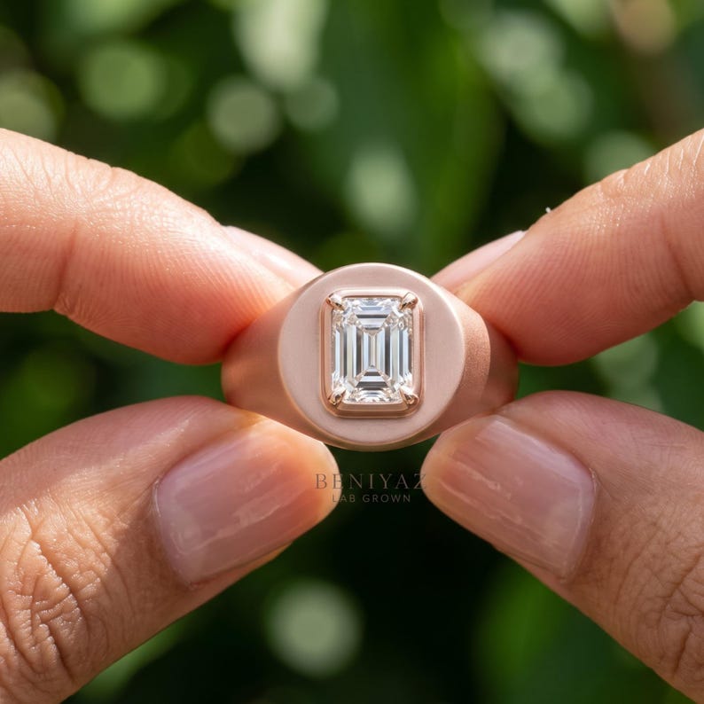 Rose gold emerald cut diamond signet ring held between fingers, showing the vertical center stone and polished signet face.