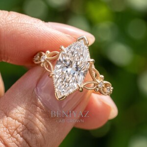 May include: A gold ring featuring a large marquise-cut center stone held by prongs. The band displays an intricate filigree design with small round accent stones embedded along the sides. The text BENIYAZ LAB GROWN is visible on the image.