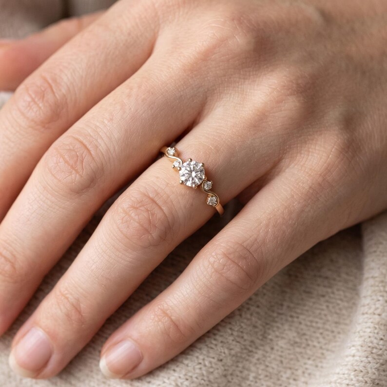 K&ouml;nnte beinhalten: Ein Ring aus Ros&eacute;gold mit einem zentralen, rund geschliffenen wei&szlig;en Edelstein in einer Krappenfassung, flankiert von zwei kleineren runden wei&szlig;en Edelsteinen auf jeder Seite, auf einer zarten, leicht geschwungenen Ringschiene.