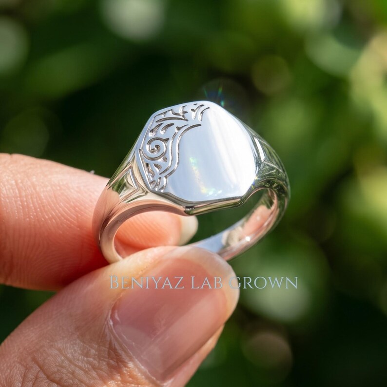 Close up sterling silver oval signet ring held by hand with smooth personalized face, decorative openwork shoulders and tapered shank