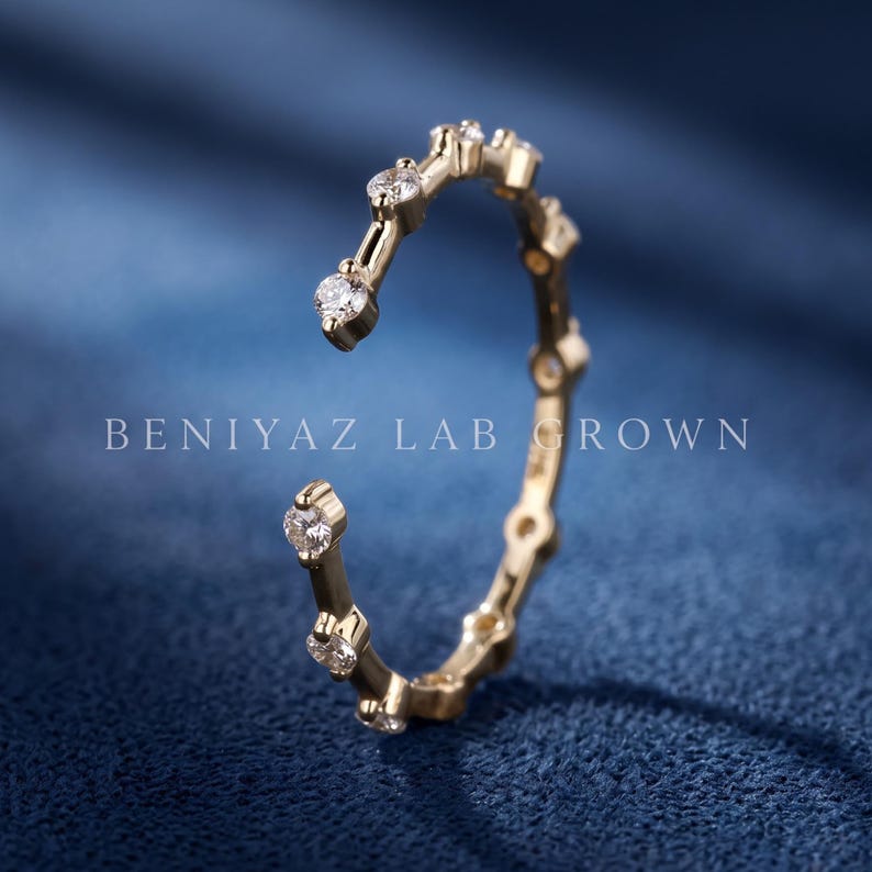 May include: A gold open-style ring featuring a series of round, clear, lab-grown stones set along the band. The text BENIYAZ LAB GROWN is visible across the center of the image. The ring has a polished finish and a circular, incomplete band design.