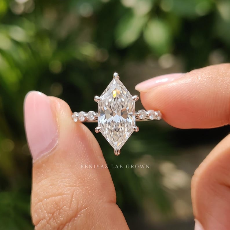 Dutch marquise solitaire engagement ring held between fingertips against a green blurred background, highlighting the distinctive elongated center diamond and delicate side stone details.