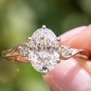 May include: A rose gold ring featuring a large oval-cut centre stone held by six prongs. The band has a curved, bypass-style design with a small round-cut stone set into each side of the shank, creating a symmetrical and elegant appearance.