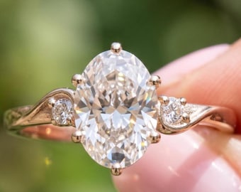 Puede incluir: Un anillo de oro rosa con una gran piedra central de corte ovalado sujeta por seis garras. La banda tiene un diseño curvo de estilo bypass con una pequeña piedra de corte redondo engastada a cada lado, creando un aspecto simétrico.