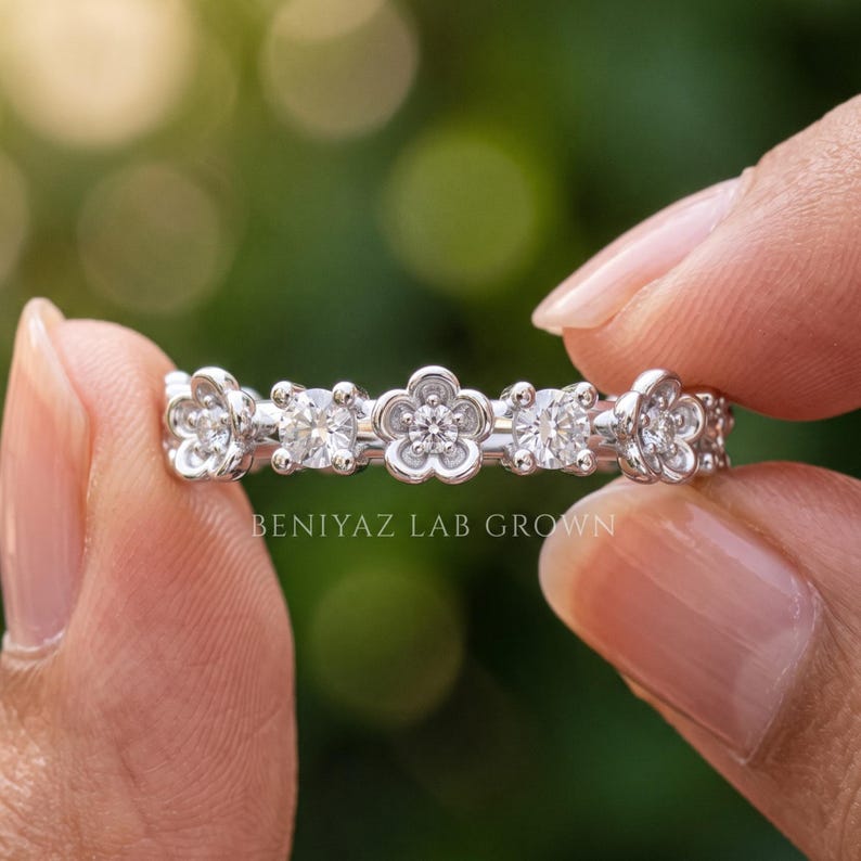 May include: A silver-toned band ring featuring a repeating pattern of three flower-shaped settings and two round-cut stones. The metal is polished and reflective. The text BENIYAZ LAB GROWN is printed in white across the center of the ring band.