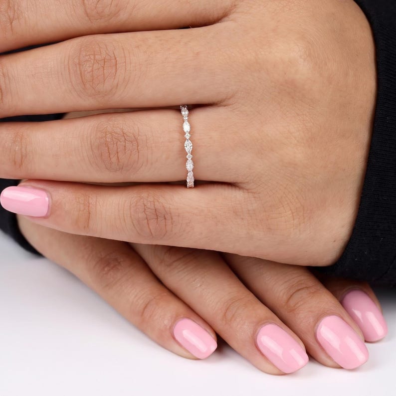 Close-up of a woman's hand displaying a thin lab grown diamond wedding band with pale pink nails and dark clothing. Marquise shaped diamonds and smaller round accents create a sparkling pattern across the upper section of the ring. The simple polished band gives this dainty half eternity design a refined appearance suitable for daily wear.