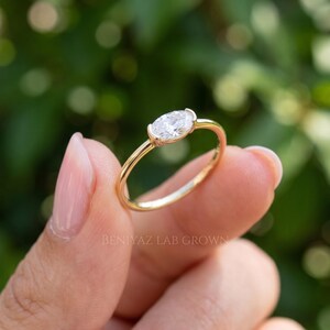 Three-quarter close-up of a horizontal oval diamond ring resting between fingertips. The sparkling oval gemstone is enclosed in a smooth yellow gold bezel, with a slender band and soft green outdoor background.