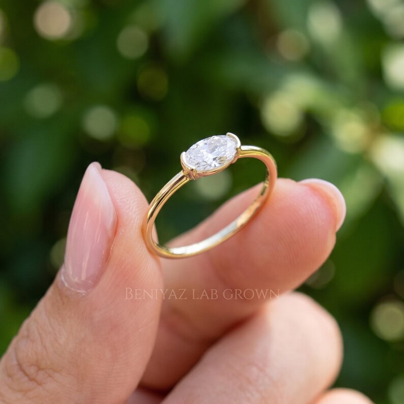 Three-quarter close-up of a horizontal oval diamond ring resting between fingertips. The sparkling oval gemstone is enclosed in a smooth yellow gold bezel, with a slender band and soft green outdoor background.