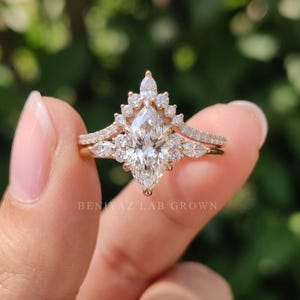 Three-quarter view of a marquise engagement ring and curved wedding band held between manicured fingertips. The pointed center diamond, clustered accent stones and coordinated rose gold tone bands are visible against a soft green garden background.