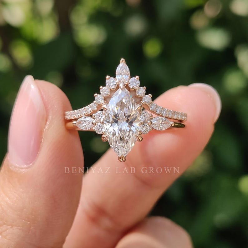 Three-quarter view of a marquise engagement ring and curved wedding band held between manicured fingertips. The pointed center diamond, clustered accent stones and coordinated rose gold tone bands are visible against a soft green garden background.