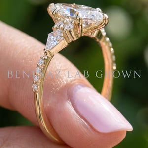 Side profile of a marquise diamond engagement ring held upright between manicured fingertips. The elevated center diamond, detailed prong setting and small side stones are visible above the slender yellow gold tone shank.