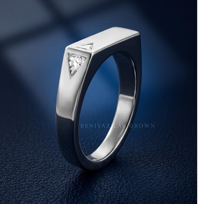 Puede incluir: Un anillo de tono plateado con una superficie superior plana y rectangular que presenta dos peque&ntilde;as piedras transparentes de corte triangular incrustadas verticalmente. El aro tiene un acabado liso y pulido, con el texto BENIYAZ LAB GROWN visible.