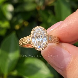 Close-up of an oval center diamond ring held gently between two fingers outdoors. The image shows the gemstone's brilliant facets, surrounding accents and detailed yellow gold tone ring design.