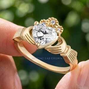 Side angle of a yellow gold Claddagh ring held between fingers, showing the raised heart diamond setting and sculpted shoulder details.