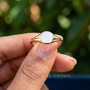 Close up round signet ring held between fingers showing smooth blank circular face, polished finish and delicate tapered band