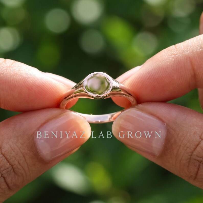Close up side view of round signet ring held between fingers showing blank polished face and slim tapered shoulders