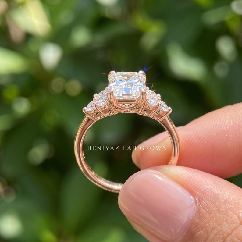 May include: A rose gold ring featuring a large rectangular radiant-cut center stone flanked by two smaller round stones on each side. The band is polished rose gold, and the text BENIYAZ LAB GROWN is visible on the inner surface of the ring band.