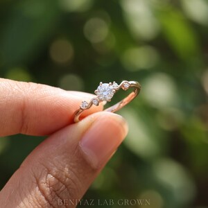 K&ouml;nnte beinhalten: Ein Ring aus Ros&eacute;gold mit einem zentralen runden Brillantschliff-Stein, der von zwei kleineren Steinen auf jeder Seite an einer geschwungenen Ringschiene flankiert wird. Der Text BENIYAZ LAB GROWN steht unten im Bild.