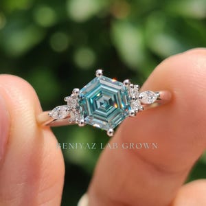 Close up blue moissanite ring featuring hexagon step cut center stone, polished prong setting and sparkling side stone cluster.