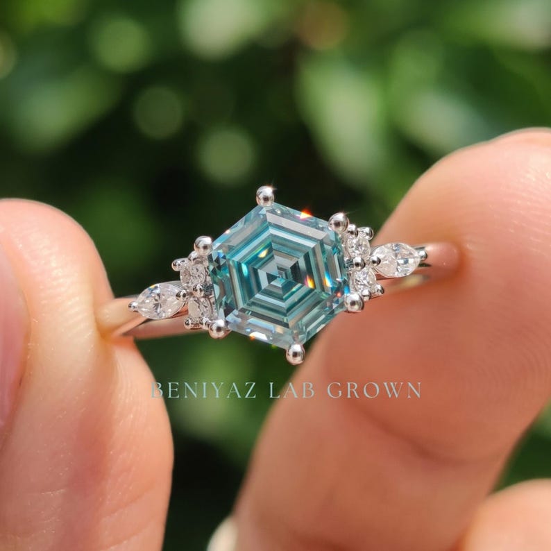 Close up blue moissanite ring featuring hexagon step cut center stone, polished prong setting and sparkling side stone cluster.