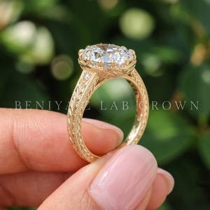 An oval diamond engagement ring photographed at a slight angle between fingertips. The image highlights the oval center stone, intricate side detailing and warm gold tone metal in natural daylight.