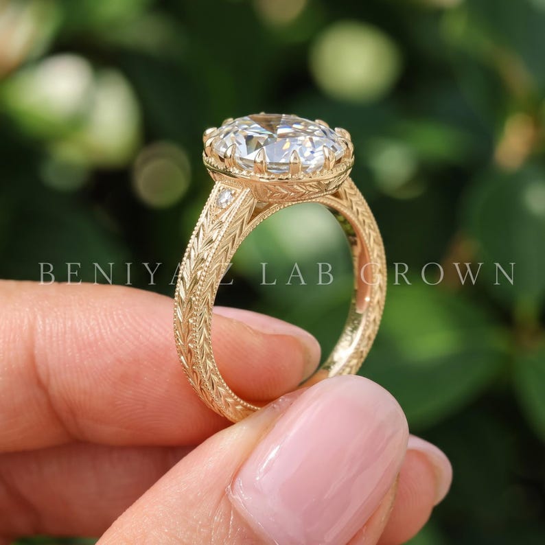 An oval diamond engagement ring photographed at a slight angle between fingertips. The image highlights the oval center stone, intricate side detailing and warm gold tone metal in natural daylight.