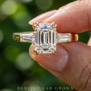 Close up three stone emerald cut lab grown diamond ring held between fingers showing step cut center and symmetrical tapered side diamonds