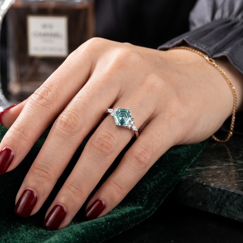 Moissanite engagement ring worn on hand, featuring a cyan blue hexagon center stone with marquise and round accent stones.