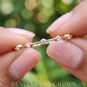 Macro photograph of a marquise and round moissanite band held horizontally between fingertips. The alternating sparkling gemstones and fine polished metal design are visible against a soft green background.