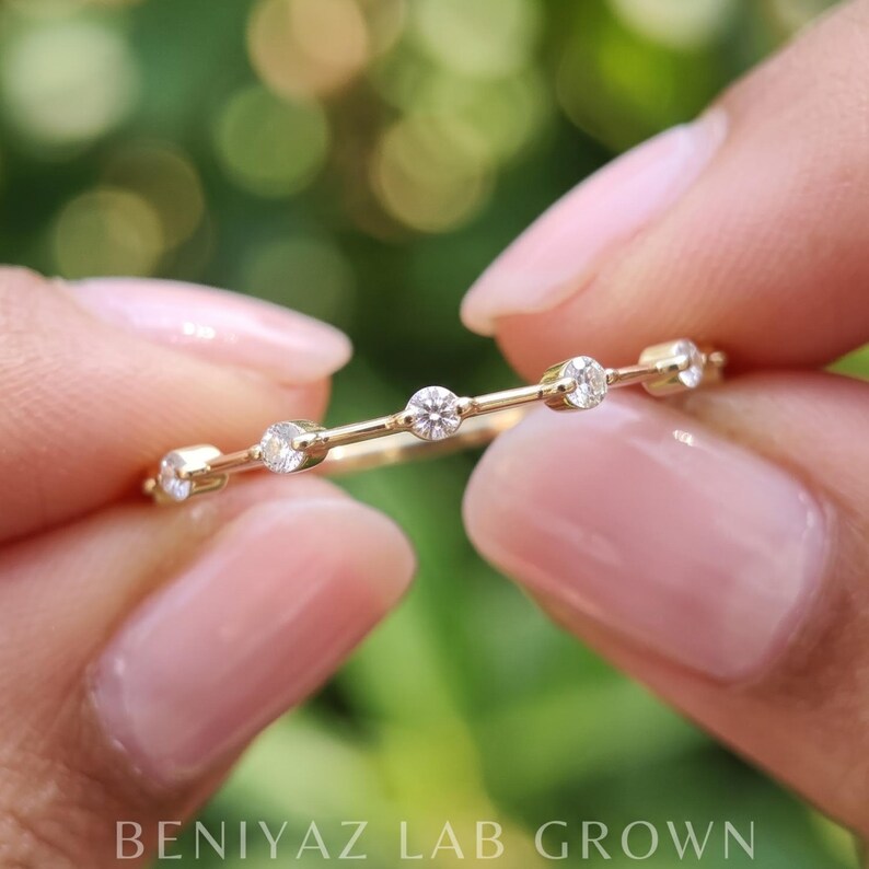 Macro photograph of a marquise and round moissanite band held horizontally between fingertips. The alternating sparkling gemstones and fine polished metal design are visible against a soft green background.