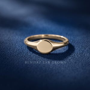Round signet ring on blue background with blank polished circular face, slim tapered shoulders and yellow gold tone finish