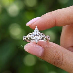 Side-angle view of a kite shaped diamond engagement ring balanced between fingertips against blurred greenery. The elevated pointed diamond center, sparkling side accents and polished gold tone infinity design are visible in natural daylight.