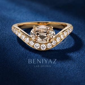 Macro side-angle photo of an oval cut lab grown diamond ring held between fingers, highlighting the yellow gold setting, center stone and pavé diamond bypass band.